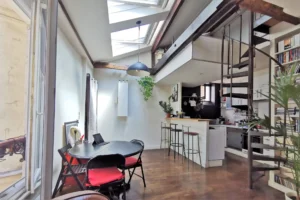 Séjour lumineux d’un loft à vendre à Paris 18, Marché de l’Olive, avec cuisine ouverte, verrière et poutres apparentes sous verrière.