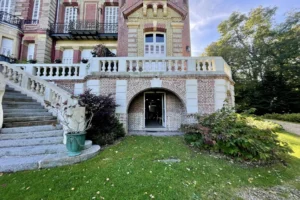 Entrée du château de Villers-sur-Mer avec son escalier en pierre, sa voûte en briques et ses balustrades blanches dans un environnement verdoyant du Calvados.