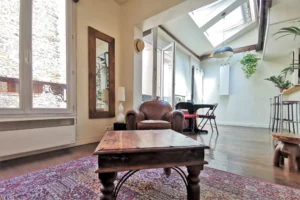 Espace salon d’un loft à vendre à Paris 18, Marché de l’Olive, avec fauteuil en cuir, miroir mural, verrière et lumière naturelle traversante.
