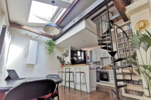 Cuisine ouverte d’un loft à vendre à Paris 18, Marché de l’Olive, avec bar en bois, escalier industriel en colimaçon et poutres apparentes sous verrière.