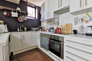 Cuisine fonctionnelle d’un loft à vendre à Paris 18, secteur Marx Dormoy – Marché de l’Olive, avec mobilier blanc, plan de travail et fenêtre sur cour intérieure.