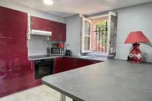 Cuisine équipée moderne de l’appartement au château de Villers-sur-Mer, avec meubles laqués rouges, plan de travail en pierre et fenêtre donnant sur le parc arboré du Calvados.