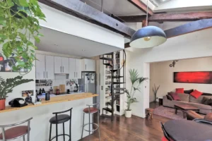 Espace de vie d’un loft à vendre à Paris 18, secteur Marx Dormoy – Marché de l’Olive, avec poutres apparentes, bar en bois et escalier design en colimaçon.