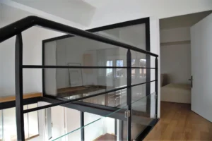 Passerelle vitrée d’un loft à Paris 19, Villa Marcel Lods, reliant la mezzanine à la chambre avec garde-corps en métal noir et verre transparent.