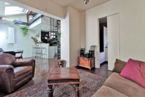 Vue du salon d’un loft à vendre à Paris 18, quartier Marx Dormoy – Marché de l’Olive, avec fauteuil en cuir, table basse en bois et ouverture sur la cuisine équipée.