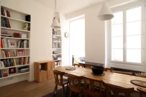 Salle à manger lumineuse d’un appartement 3 pièces situé rue du Faubourg Saint-Denis à Paris 10, avec grande table en bois, bibliothèque intégrée et fenêtre double vitrage.