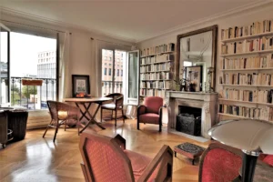 Séjour lumineux avec bibliothèques murales, cheminée en marbre, parquet en point de Hongrie et larges fenêtres offrant une vue dégagée sur le square Alban Satragne, dans un appartement situé rue du Faubourg Saint-Denis à Paris 10.