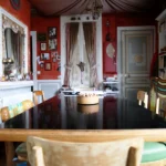 Salle à manger décorée de façon éclectique avec grande table en bois, moulures au plafond, tissu tendu, murs rouges et espace de travail dans un appartement situé rue du Faubourg Saint-Denis à Paris 10.