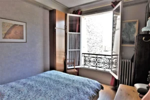 Chambre d’un duplex rue du Faubourg Saint-Denis, avec lit double, placard en bois, fenêtre ouverte sur cour intérieure et garde-corps en fer forgé.