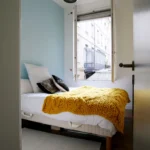 Chambre lumineuse d’un appartement situé rue du Faubourg Saint-Denis à Paris 10, avec mur bleu pastel, grande fenêtre ouverte et couverture jaune en maille épaisse.