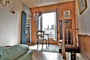 Espace nuit en mezzanine d’un duplex rue du Faubourg Saint-Denis, avec palier en bois, table près d’une fenêtre ouverte sur balcon et vue sur les toits de Paris 10.