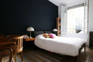 Grande chambre d’un appartement situé rue du Faubourg Saint-Denis à Paris 10, avec mur bleu nuit, bureau en bois, lit double et grande fenêtre ouverte sur cour.