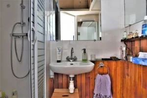 Salle de douche d’un duplex rue du Faubourg Saint-Denis, avec cabine de douche vitrée, petit lavabo, rangement en bois et miroir mural.