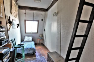 Petite chambre sous combles d’un duplex rue du Faubourg Saint-Denis, avec lit simple, poutres apparentes, fenêtre mansardée et sol en tomettes.