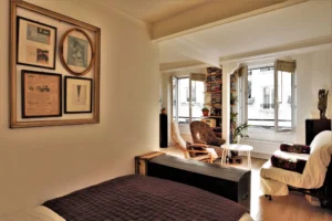 Espace nuit ouvert sur le séjour d’un appartement rue Poissonnière Paris 2, avec lit, fauteuils vintage, grande lumière naturelle et fenêtres sur cour.