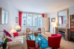 Salon lumineux avec grandes fenêtres donnant sur cour intérieure, cheminée en marbre, miroir doré et fauteuils rouges dans un appartement haussmannien à Paris 16.