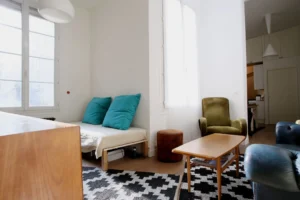 Séjour lumineux d’un appartement situé rue du Faubourg Saint-Denis à Paris 10, avec banquette, fauteuil vert, tapis graphique noir et blanc et grande fenêtre double vitrage.