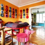 Vue d’une chambre-séjour colorée avec parquet en point de Hongrie, lit intégré avec rangements, décor mural de coussins ronds et ouverture vers une salle à manger lumineuse dans un appartement rue du Faubourg Saint-Denis à Paris 10.