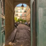 Vue depuis le porche d’entrée du Passage Delanos à Paris 10, donnant sur une cour pavée entourée d’immeubles aux volets verts, avec une lumière traversante et une ambiance d’atelier parisien.
