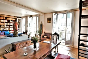 Salle à manger lumineuse d’un duplex rue du Faubourg Saint-Denis, avec grande table en bois, fauteuils sculptés, bibliothèque et ouvertures sur balcon dans un appartement ancien à Paris 10.