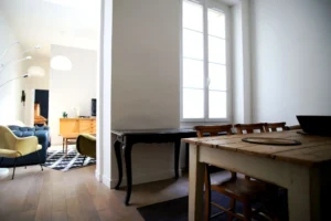 Vue ouverte entre le séjour et la salle à manger d’un appartement rue du Faubourg Saint-Denis à Paris 10, avec fauteuils vintage, parquet et grande fenêtre.