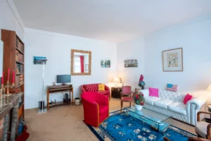 Séjour spacieux dans un appartement haussmannien à Paris 16, avec canapé en cuir blanc, fauteuils rouges, tapis bleu, bibliothèque ancienne et miroir doré.