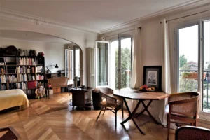 Séjour spacieux avec bibliothèque, coin bureau, parquet en point de Hongrie et grandes fenêtres ouvertes offrant une vue dégagée sur le square Alban Satragne depuis la rue du Faubourg Saint-Denis à Paris 10.