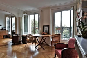 Coin repas lumineux avec table en bois et chaises en rotin, parquet en point de Hongrie et larges fenêtres ouvertes offrant une vue dégagée sur le square Alban Satragne depuis la rue du Faubourg Saint-Denis à Paris 10.
