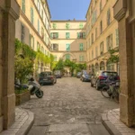 Cour pavée du Passage Delanos à Paris 10, encadrée d’immeubles aux volets verts et d’arcades végétalisées, avec voitures, vélos et lumière naturelle.