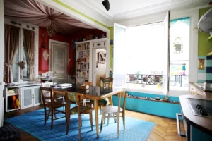 Salle à manger lumineuse avec table en bois, parquet en point de Hongrie, grande fenêtre ouverte, éléments décoratifs colorés et cuisine attenante dans un appartement situé rue du Faubourg Saint-Denis à Paris 10.