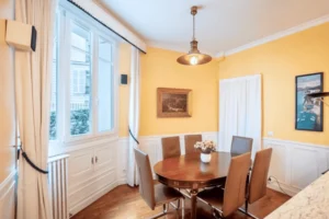 Salle à manger lumineuse avec murs jaunes, grande fenêtre donnant sur cour, table ronde en bois et chaises contemporaines dans un appartement situé rue de la Pompe à Paris 16.