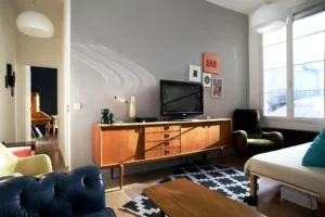 Séjour lumineux d’un appartement rue du Faubourg Saint-Denis à Paris 10, avec buffet vintage en bois, fauteuils rétro, grande fenêtre et décoration graphique.