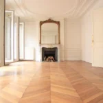 Salon haussmannien avec parquet point de Hongrie, grande cheminée en marbre surmontée d’un miroir ancien et moulures décoratives, dans un appartement rue Beaurepaire à Paris 10.
