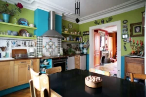 Cuisine ouverte décorée de façon colorée avec crédence en carreaux, rangements en bois, étagères ouvertes, hotte inox et vue sur le séjour avec cheminée dans un appartement situé rue du Faubourg Saint-Denis à Paris 10.