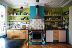 Cuisine colorée avec hotte inox centrale, crédence en mosaïque, rangements en bois, étagères ouvertes et nombreux éléments décoratifs dans un appartement situé rue du Faubourg Saint-Denis à Paris 10.
