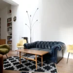 Salon d’un appartement situé rue du Faubourg Saint-Denis à Paris 10, avec canapé Chesterfield bleu, fauteuils vintage, bibliothèque intégrée et tapis graphique noir et blanc.