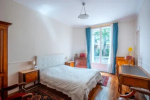 Chambre parentale avec lit double, parquet en bois, mobilier ancien et grande fenêtre donnant sur une cour arborée, dans un appartement rue de la Pompe à Paris 16.