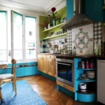 Cuisine lumineuse avec grande fenêtre, rangements en bois, hotte inox, crédence en mosaïque colorée et étagères ouvertes dans un appartement situé rue du Faubourg Saint-Denis à Paris 10.