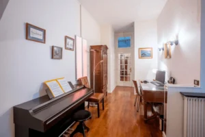 Espace bureau avec piano, parquet en bois et éclairage mural, donnant sur un couloir menant à une porte vitrée, dans un appartement rue de la Pompe à Paris 16.