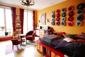 Grande chambre-séjour lumineuse avec balcon filant, décor coloré, mur orangé, lit avec rangements intégrés, fauteuils et bibliothèque dans un appartement situé rue du Faubourg Saint-Denis à Paris 10.