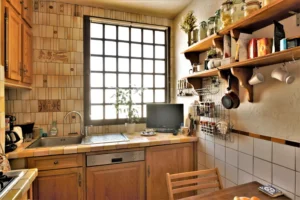 Cuisine chaleureuse d’un duplex rue du Faubourg Saint-Denis, avec carrelage ancien, meubles en bois, étagères ouvertes et fenêtre à grille laissant entrer la lumière.