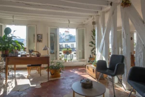 Pièce de vie lumineuse d’un appartement duplex rue d’Aboukir, avec poutres apparentes, tomettes anciennes, bureau en bois, plantes vertes et vues dégagées sur les toits parisiens.