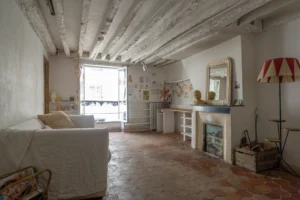 Photo du séjour d’un appartement ancien à Paris 10, rue du Faubourg Saint-Martin, avec plafond à poutres apparentes peintes en blanc, sol en tomettes, cheminée ancienne avec miroir, bureau mural et grande fenêtre ouverte sur rue.