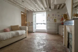 Séjour d’un appartement ancien à Paris 10, rue du Faubourg Saint-Martin, avec plafond à poutres apparentes blanchies, sol en tomettes, canapé clair, cheminée ancienne et grande fenêtre ouverte sur rue.