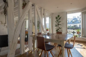 Salle à manger lumineuse d’un duplex rue d’Aboukir, avec table en bois, chaises design, poutres apparentes blanches et vue dégagée sur les toits parisiens.