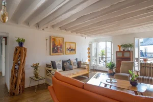 Salon lumineux d’un appartement duplex rue d’Aboukir, avec poutres apparentes, canapé en cuir, banquette décorative, meubles en bois sculpté et vue sur les toits de Paris.