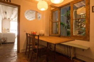 Salle à manger d’un appartement ancien à Paris 10, rue du Faubourg Saint-Martin, avec table en bois, chaises vintage, tomettes au sol, fenêtres intérieures donnant sur un patio végétalisé et suspensions lumineuses.