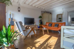 Salon lumineux d’un duplex rue d’Aboukir, avec parquet ancien, canapé en cuir orange, fauteuils vintage, table basse en bois et poutres apparentes blanches.