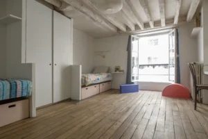 Grande chambre d’un appartement à Paris 10, rue du Faubourg Saint-Martin, avec parquet ancien, plafond à poutres apparentes blanchies, lits intégrés avec rangements et grande fenêtre donnant sur rue.