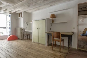 Espace nuit et bureau d’un appartement à Paris 10, rue du Faubourg Saint-Martin, avec parquet ancien, plafond à poutres apparentes blanchies, placard intégré, bureaux en bois et fenêtre donnant sur rue.